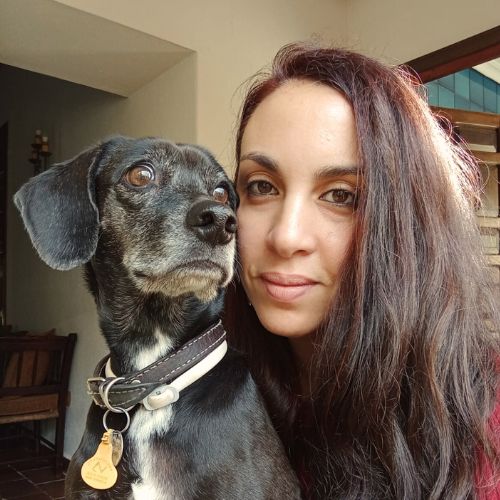 Chica morena mirando a cámara junto con una perrita negra que mira de lado. Es socia de Igualdad Animal.